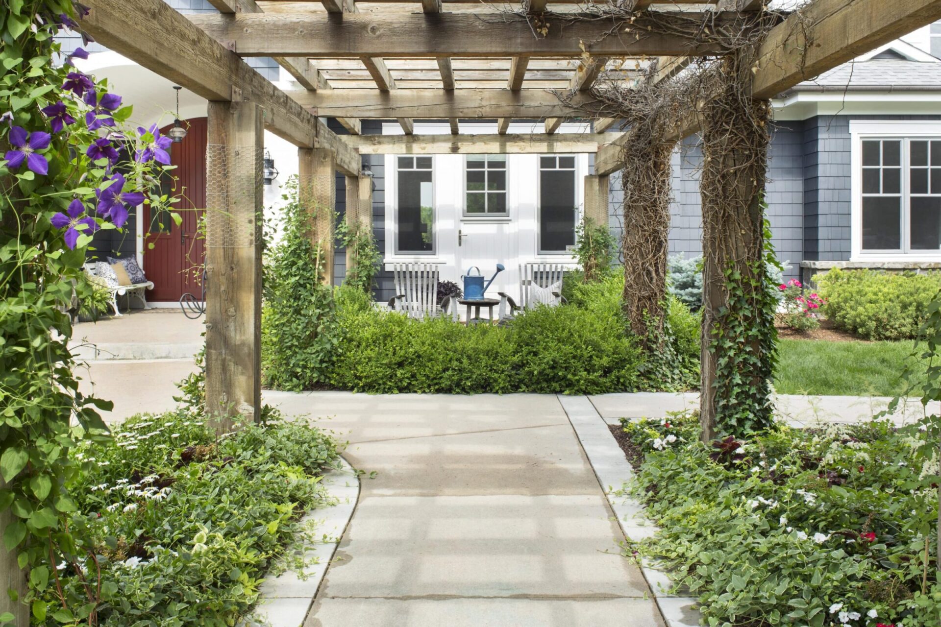 A garden path with a wooden pergola, vibrant flowers, and seating area near a house entrance with a red door.