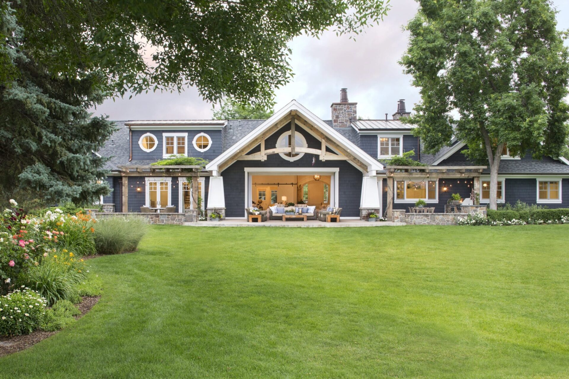 Charming house with a large green lawn, surrounded by trees and colorful flowers, showcasing a cozy outdoor seating area on the patio.