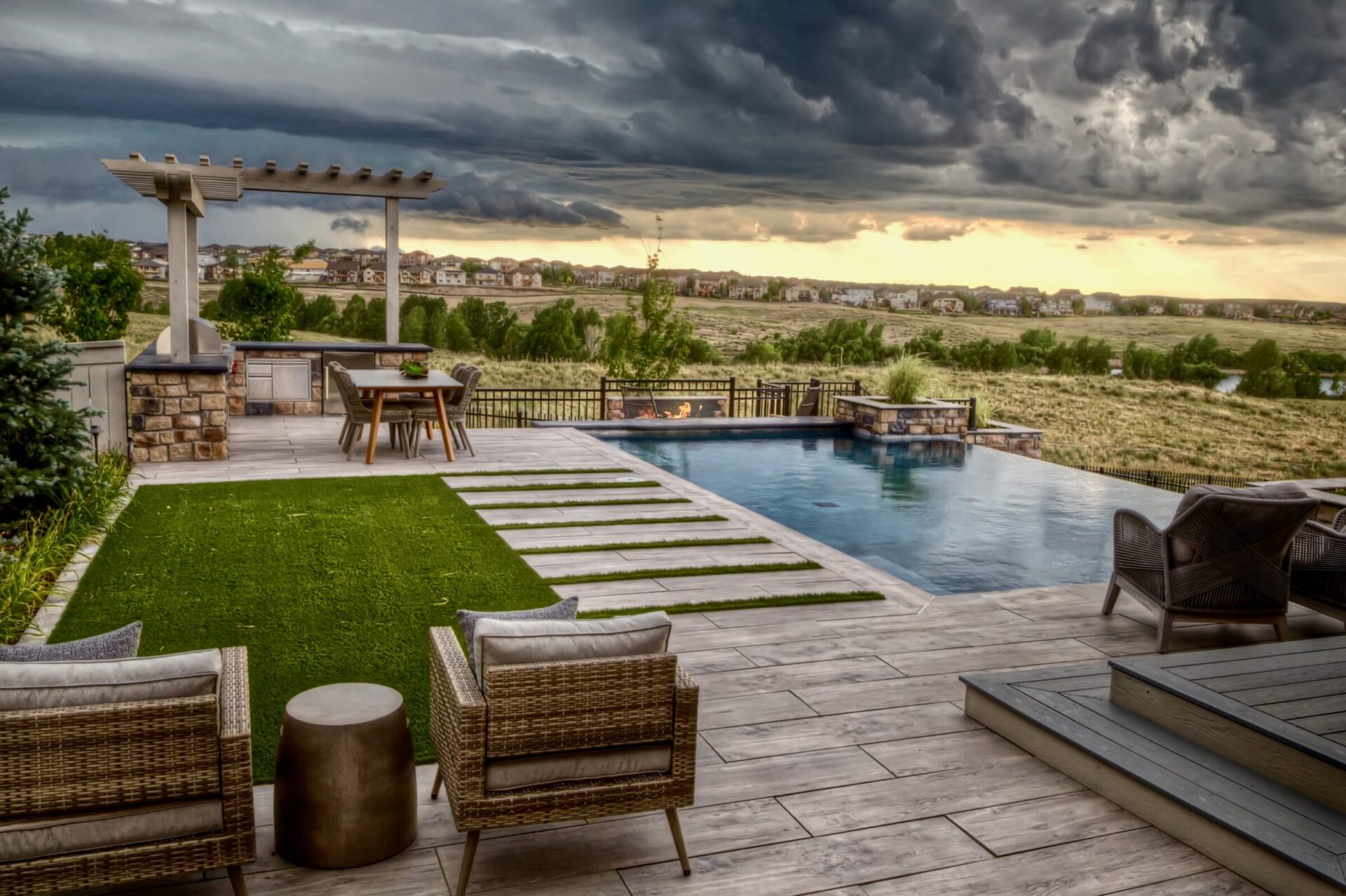 Modern patio with wicker furniture, overlooking an infinity pool and scenic landscape. Dramatic clouds create a striking backdrop over distant homes.