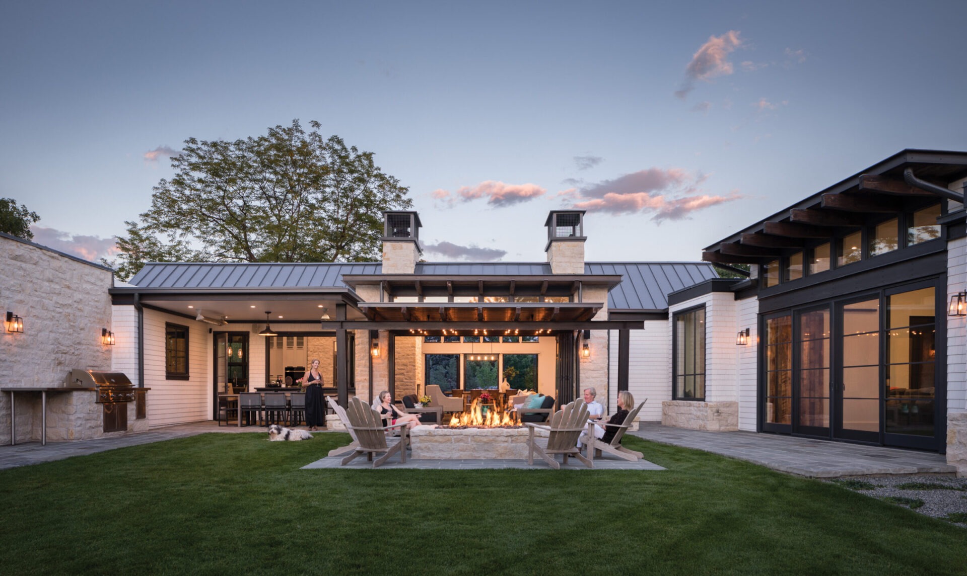 A modern house with a patio and fire pit. People seated around, enjoying an evening outdoors, surrounded by greenery and subtle lighting.