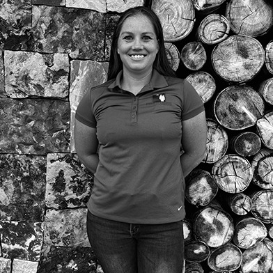 A person stands outdoors in front of a stack of cut logs and a stone wall, smiling and wearing a polo shirt.