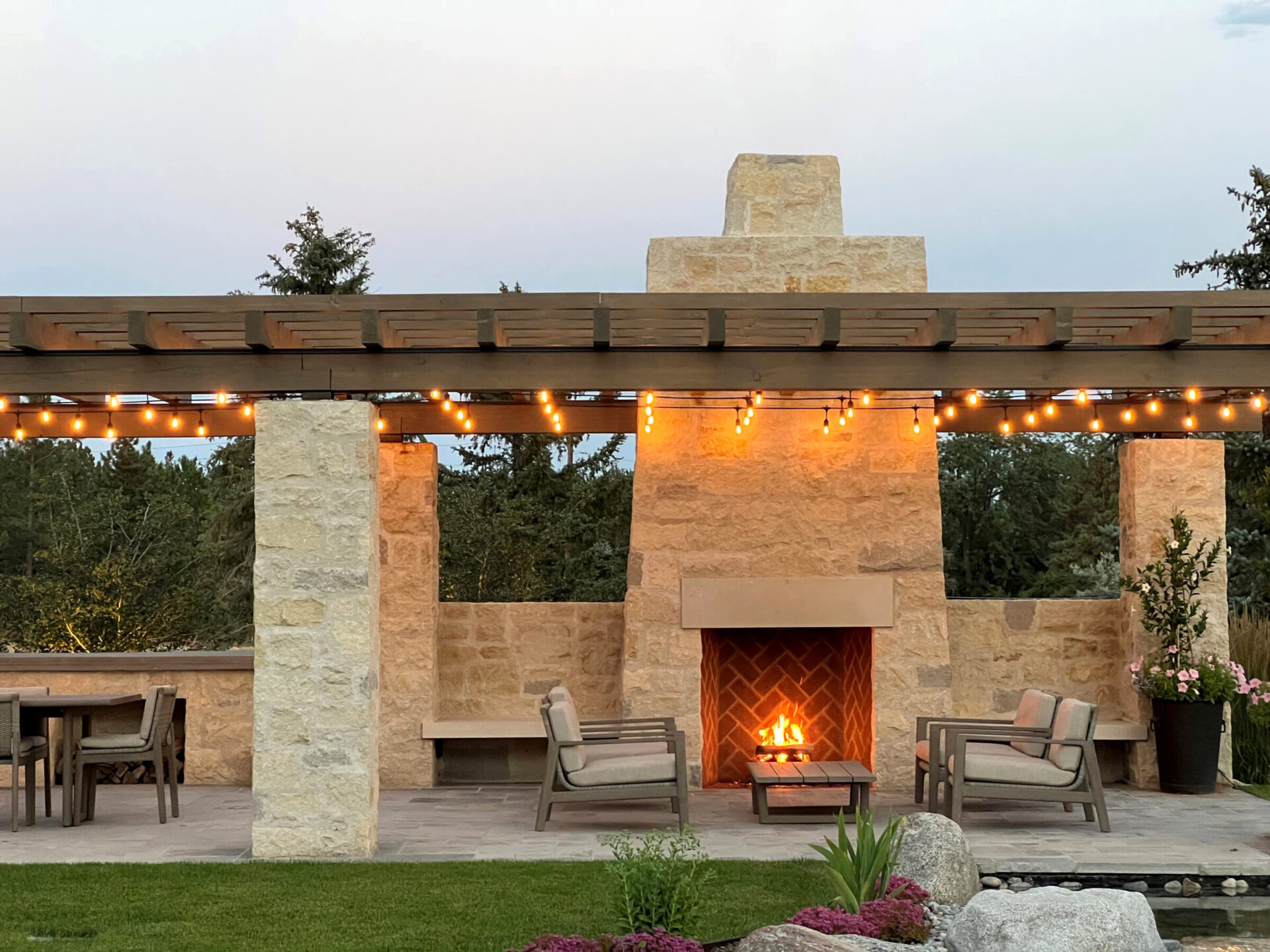 Outdoor patio with stone fireplace, string lights, and cozy seating. Surrounded by trees and garden, creating a warm, inviting atmosphere.