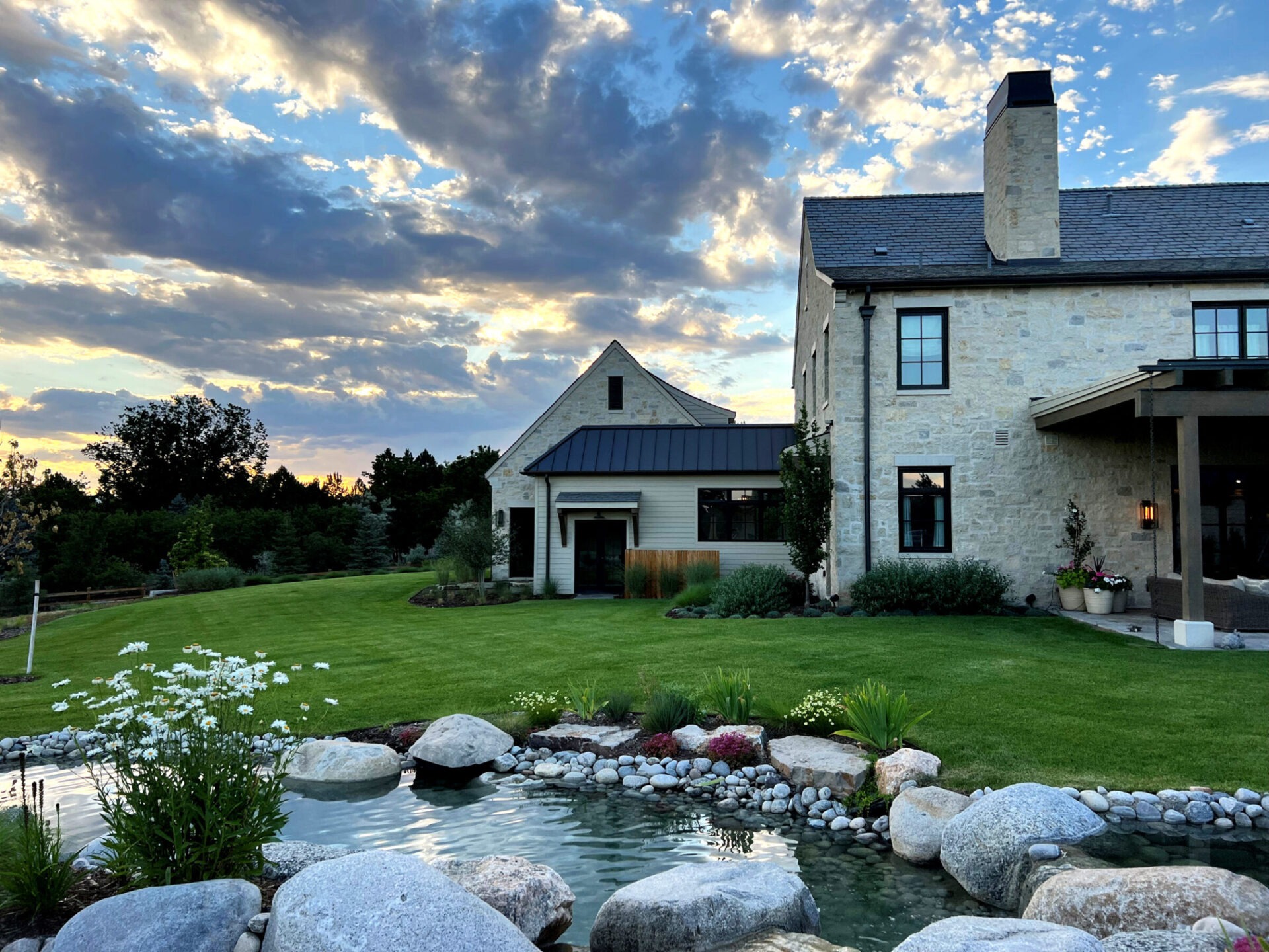 A picturesque stone house with a landscaped yard, pond, and vibrant garden, set against a dramatic sky with scattered clouds and sunset.