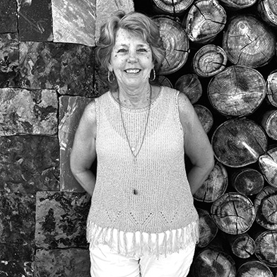 A person smiling, standing in front of a wall with stacked stones and round wooden logs, wearing a knitted top and light pants.