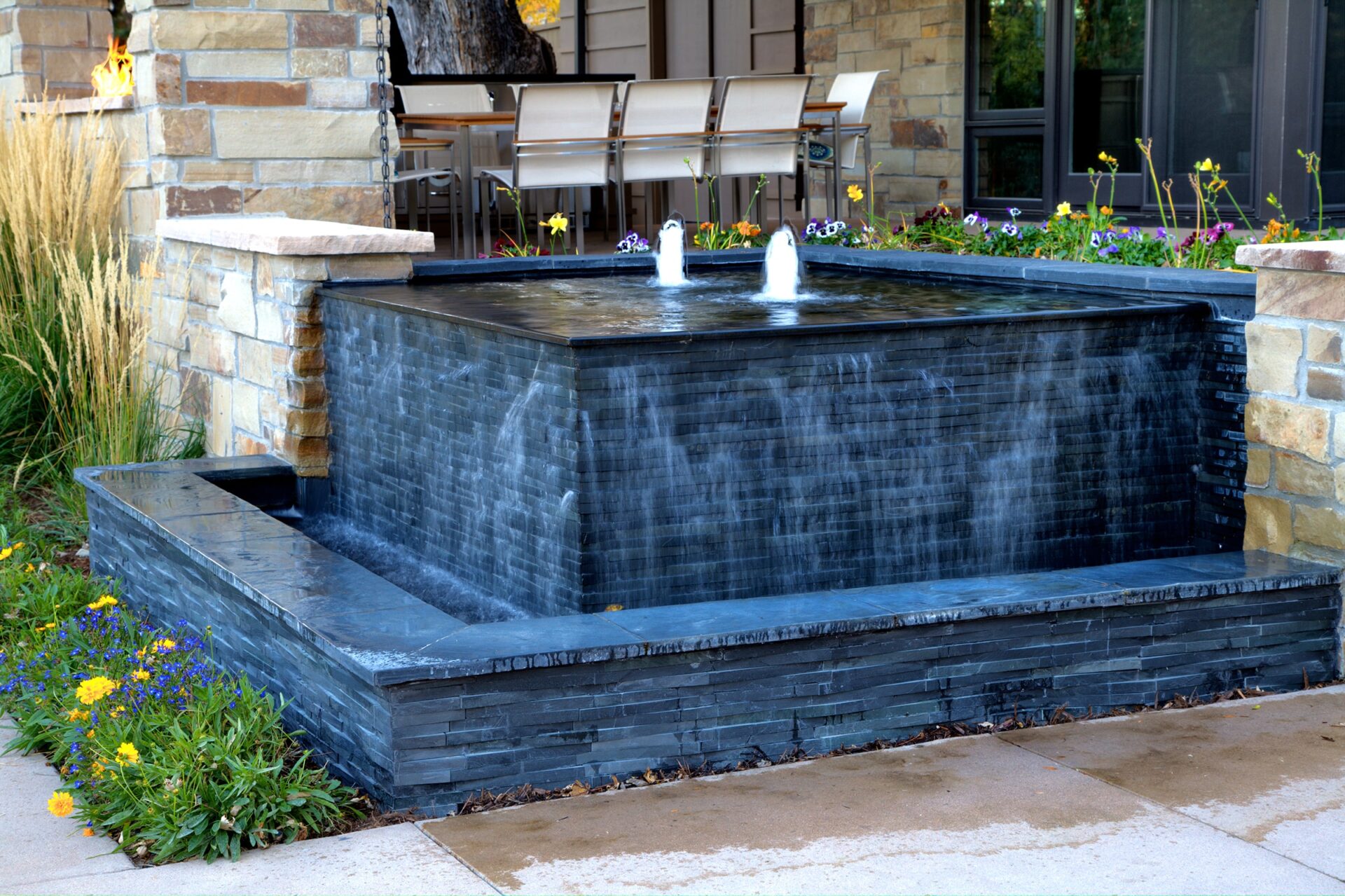 A modern stone water fountain with cascading water, surrounded by flowers and patio seating, is situated in a landscaped outdoor area.