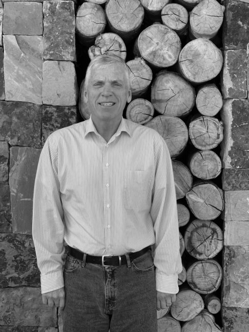 A smiling person stands in front of stacked logs and stone bricks, wearing a striped shirt and jeans, in a monochrome setting.