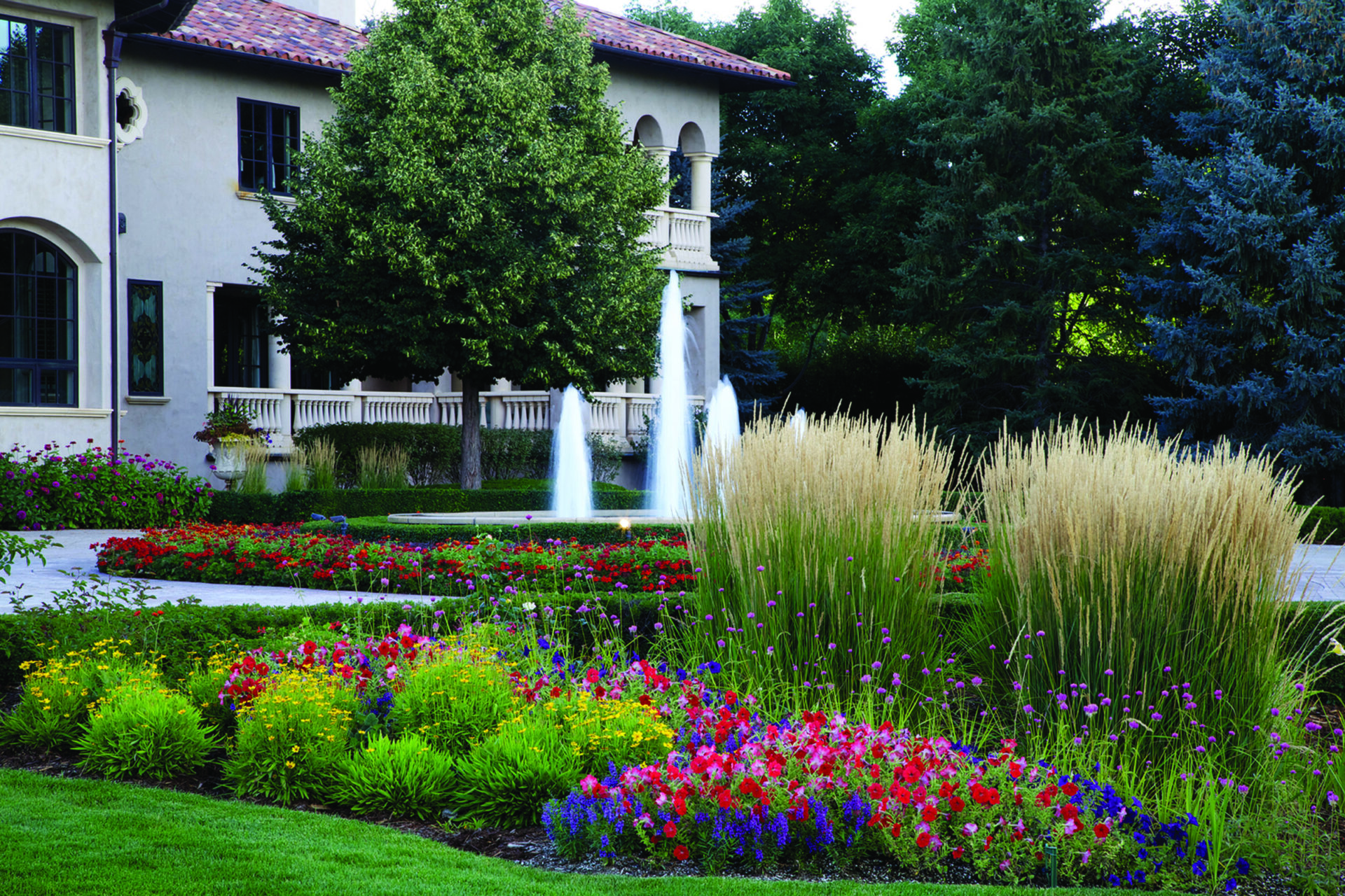 A lush garden with vibrant flowers surrounds a fountain near a Mediterranean-style building, bordered by trees, creating a tranquil atmosphere.