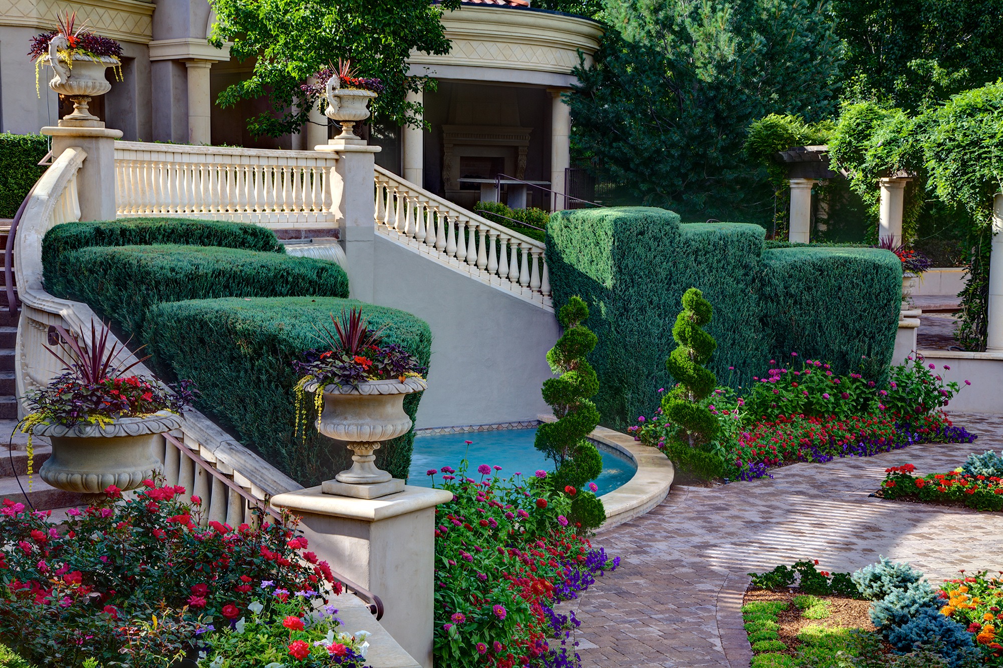 Elegant garden with manicured hedges, colorful flowers, stone pathways, and decorative urns. Stairs lead to a stately building with columns.