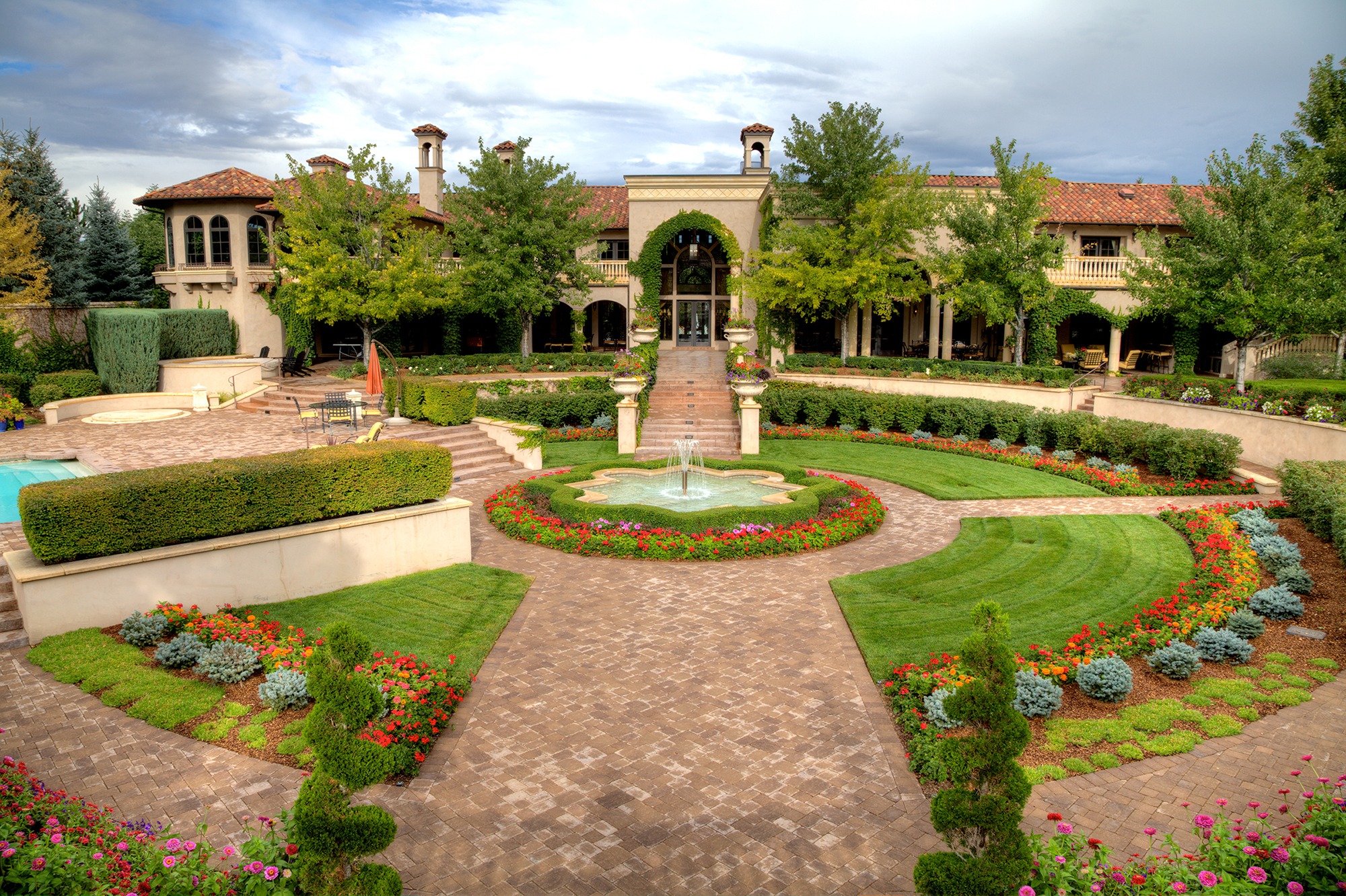 Elegant mansion with manicured gardens, vibrant flowers, a central fountain, and stone pathways, framed by lush trees and featuring a Mediterranean architectural style.