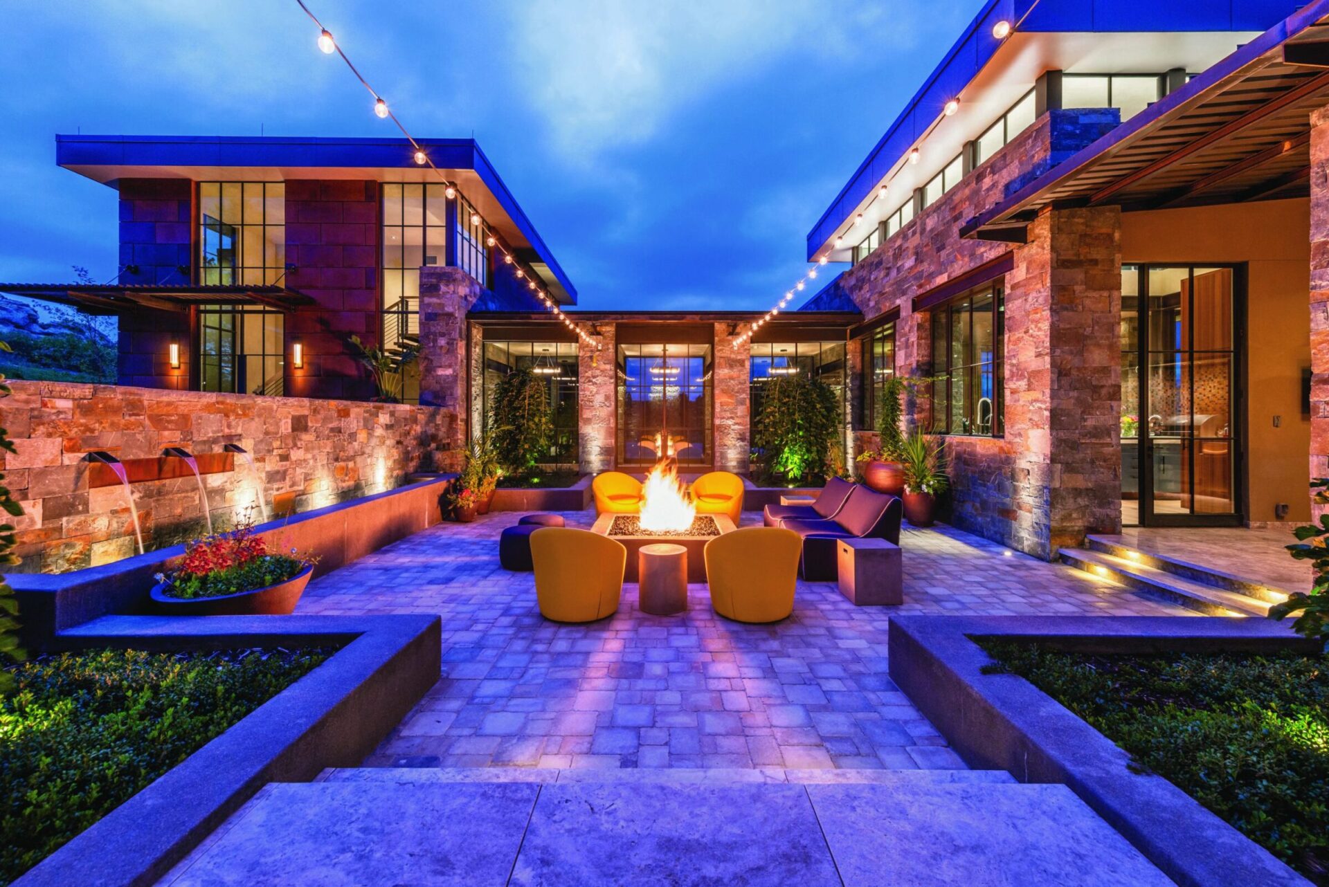 Modern outdoor patio with string lights, central fire pit, vibrant seating, and stone architecture set against a twilight sky, creating a cozy ambiance.