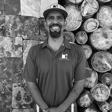 A person in a polo shirt and cap stands smiling in front of a stacked woodpile and textured stone wall, outdoors.