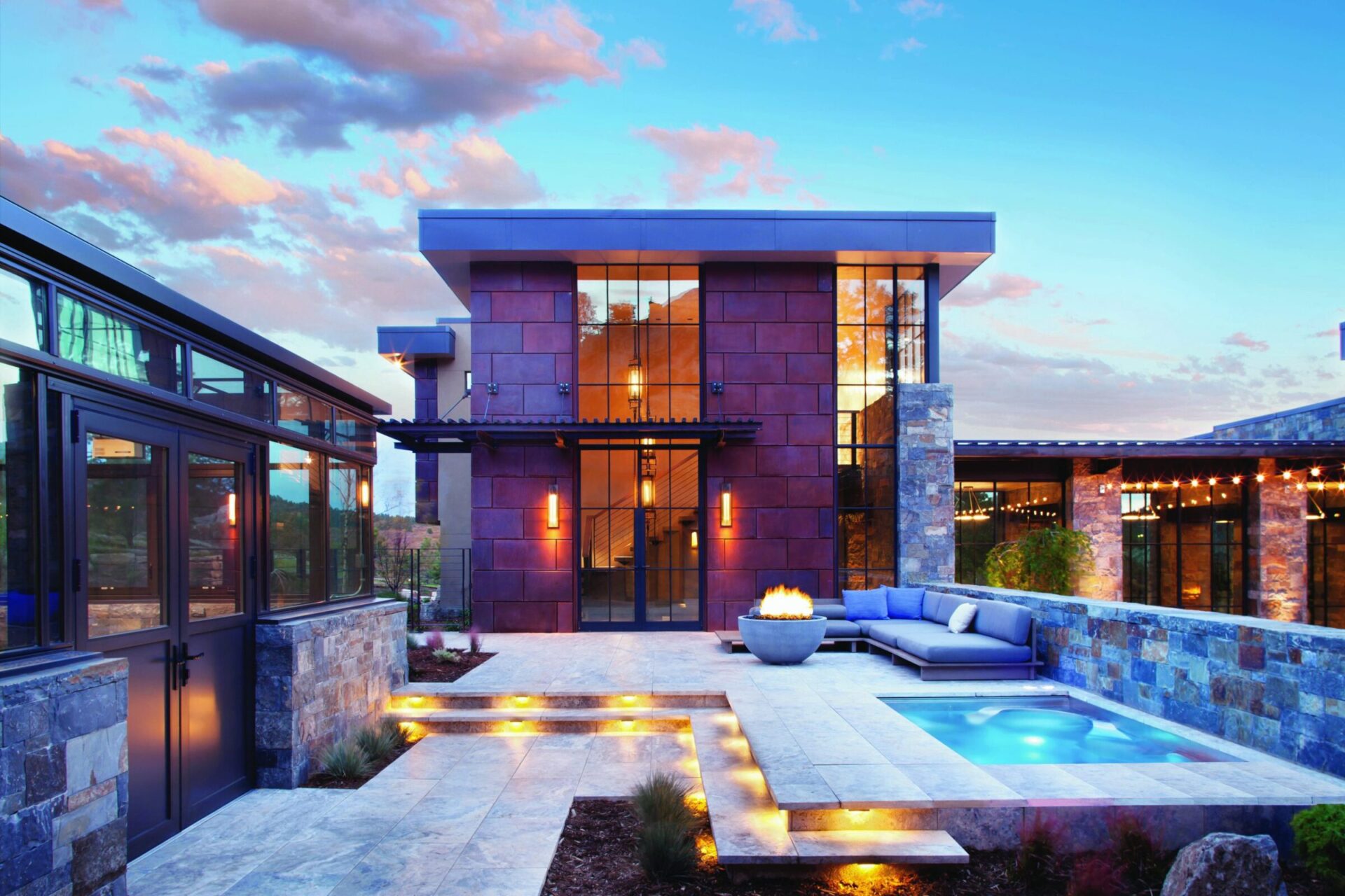 Modern home exterior with large glass windows, stone accents, and illuminated steps. Features a pool, fire pit, and patio with seating.