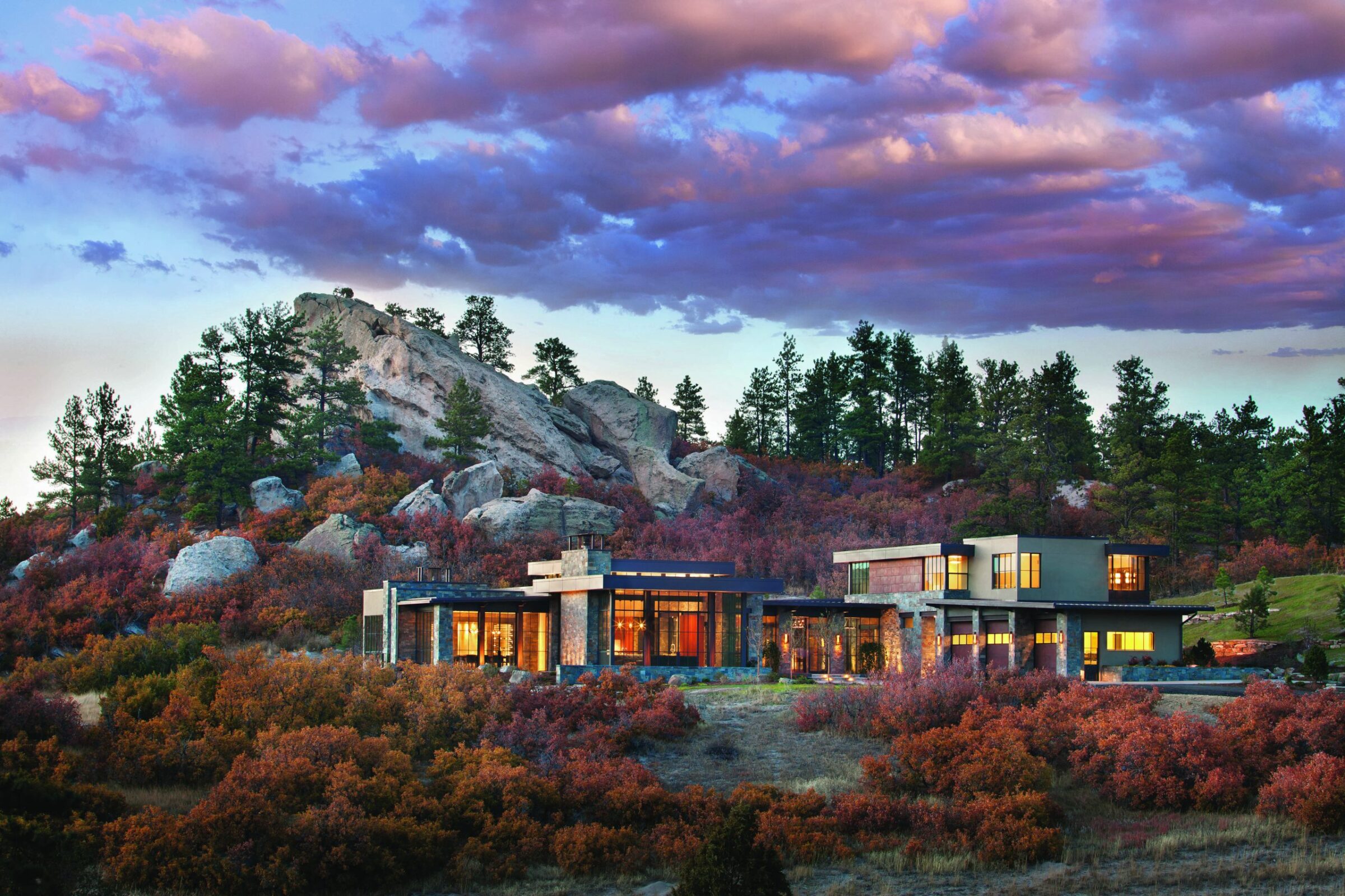 Modern house nestled in a vibrant, rocky landscape with tall trees and colorful shrubs, under a picturesque, cloud-filled sky at sunset.