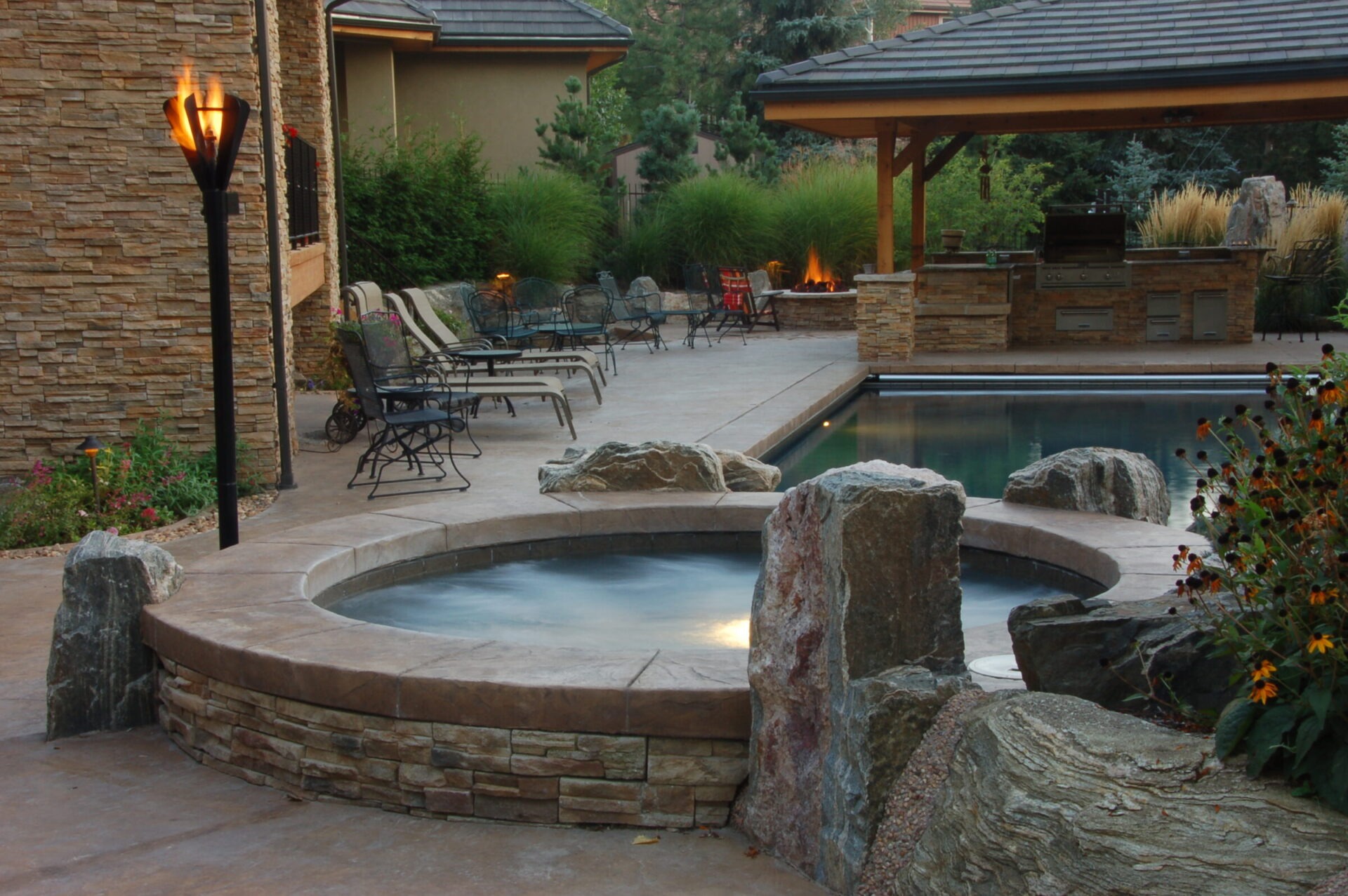 A serene backyard features a pool, chairs, stone hot tub, tiki torches, barbecue area, surrounded by plants and stone architecture.