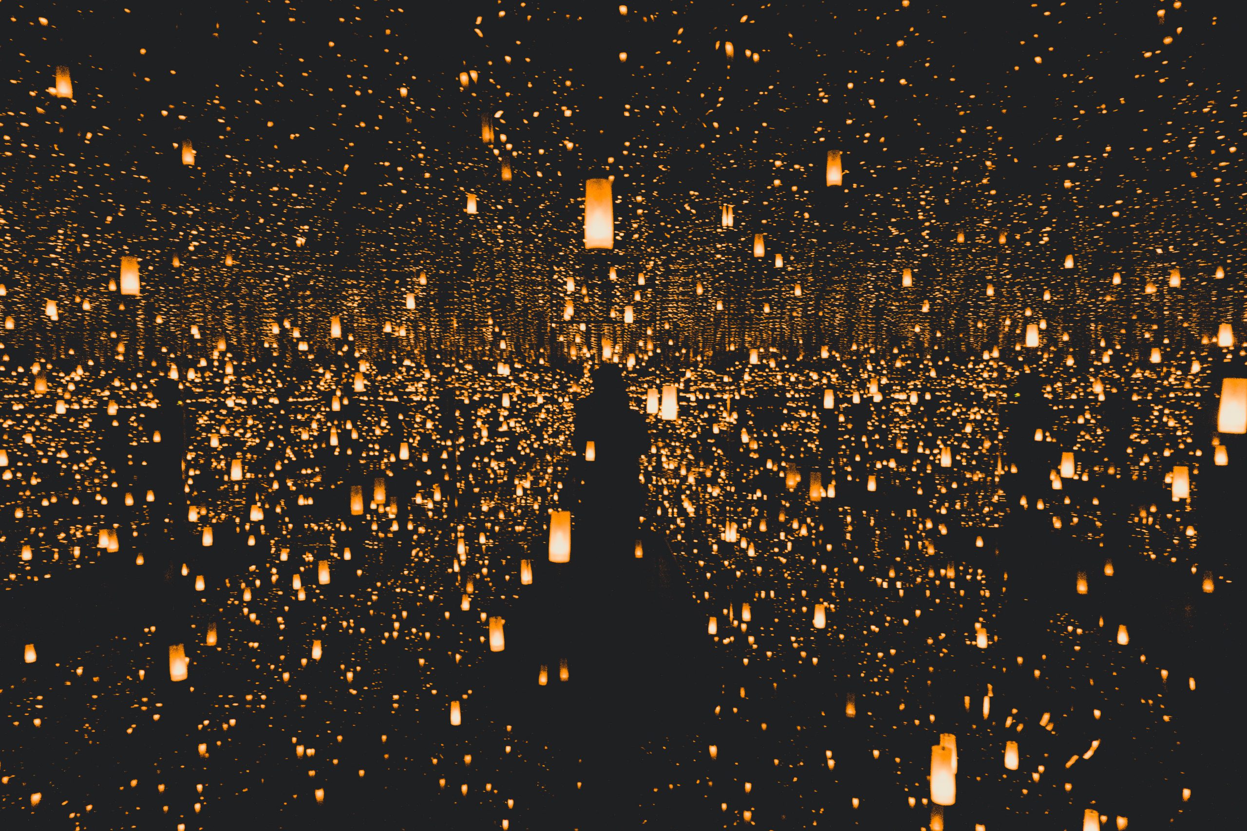 A person stands in a mirrored room filled with countless glowing, hanging lanterns, creating an illusion of infinite lights in darkness.