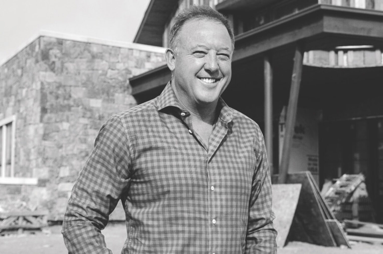 A person in a checkered shirt smiles in front of a stone building under construction, on a sunny day.