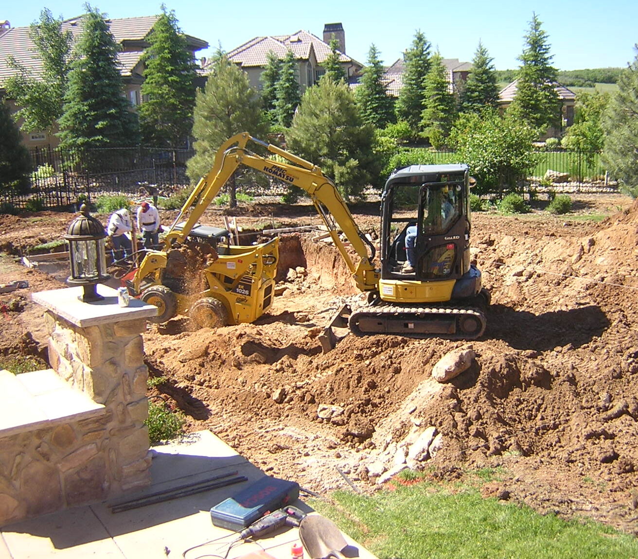 A small digger operates in a construction site, with three people nearby. Trees and residential houses are in the background.