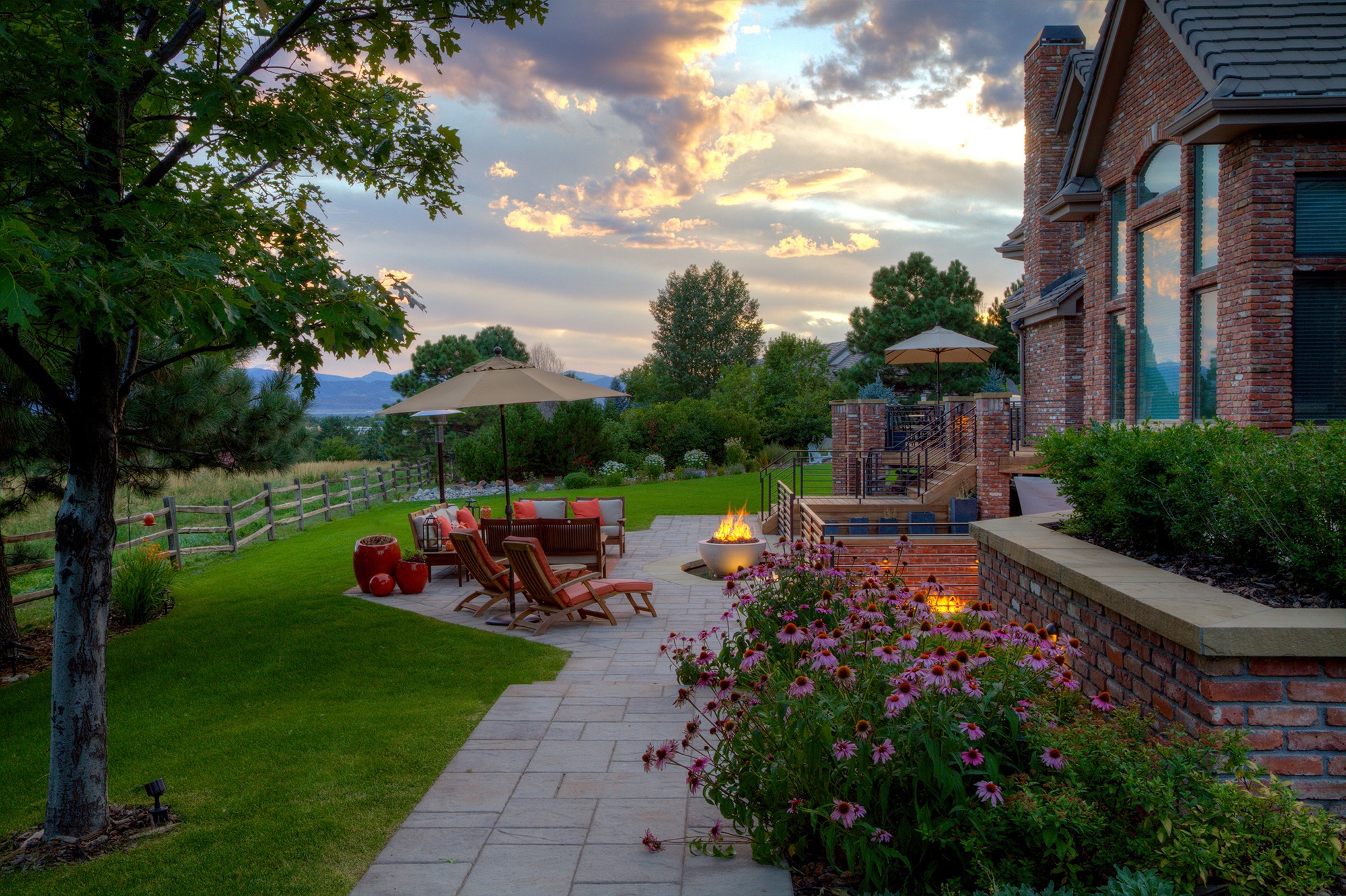 Brick house with patio featuring chairs, umbrellas, a fire pit, and vibrant flowers. Fenced lush garden under a cloudy sunset sky.