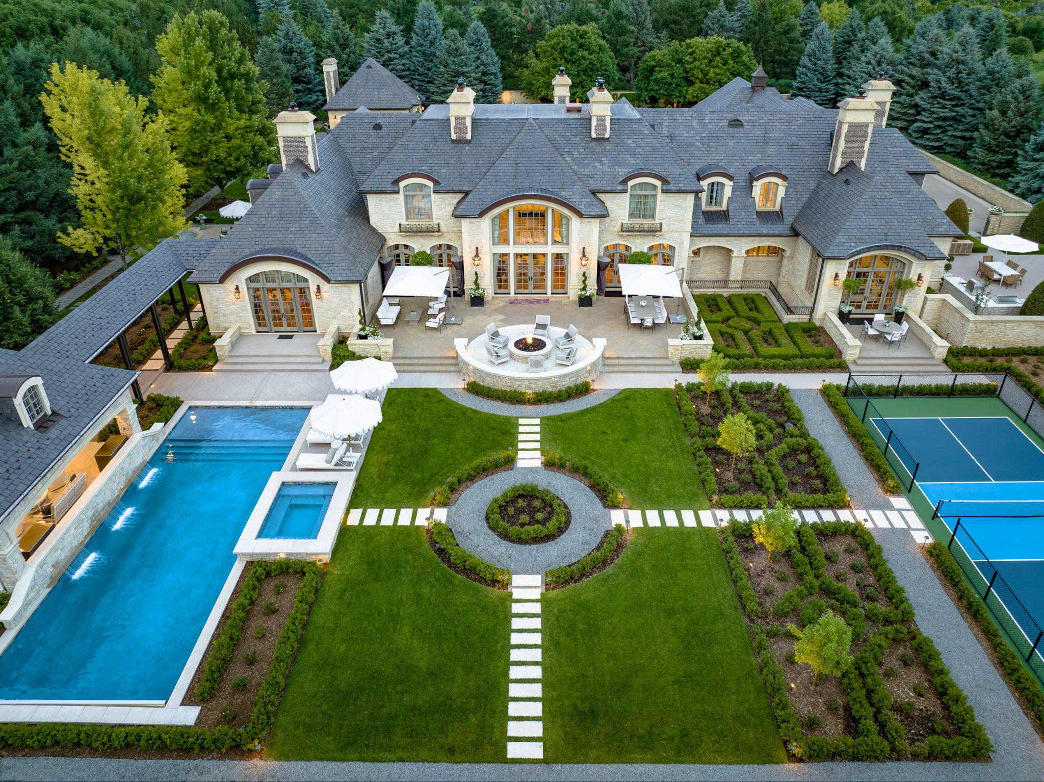Aerial view of a grand French chateau-style home with symmetrical gardens, swimming pool, patio seating, and tennis court.