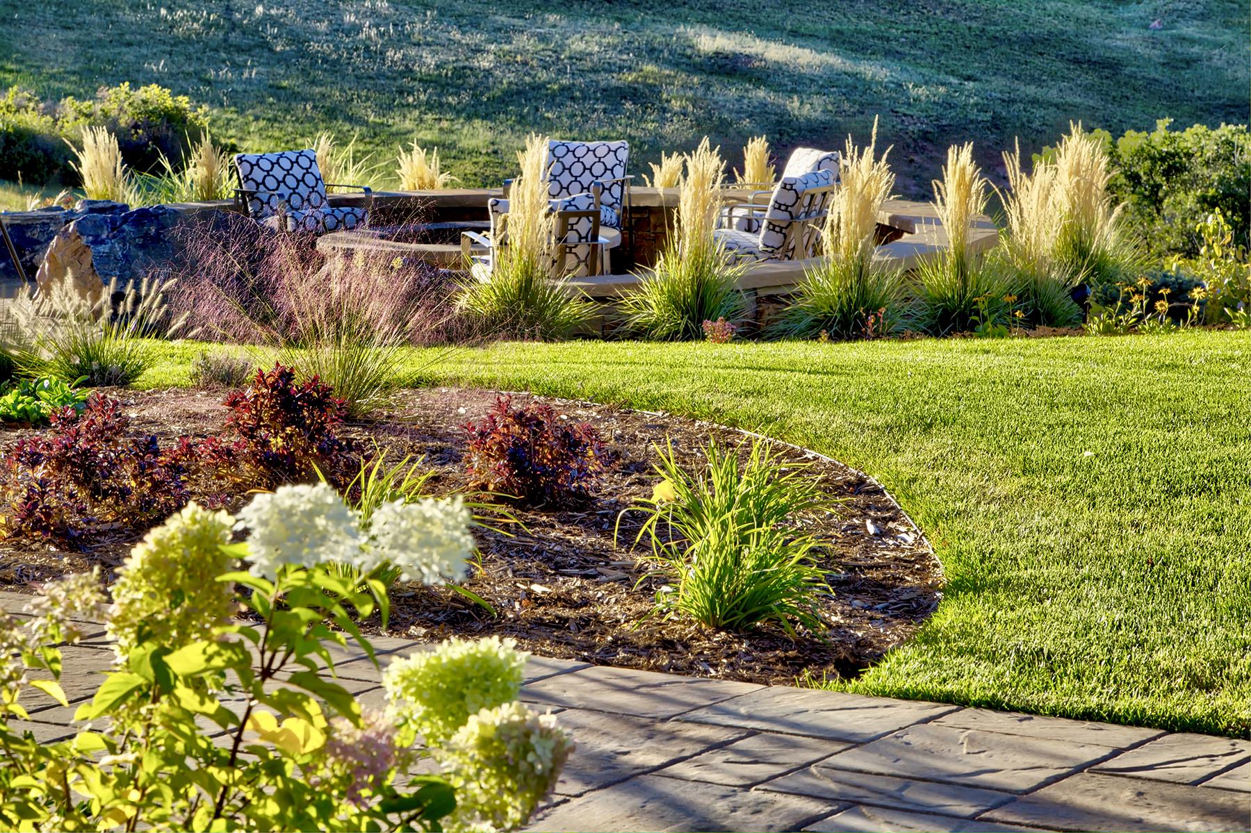 Curved landscaped garden bed with ornamental grasses and flowering plants bordering a lush green lawn and paved pathway.