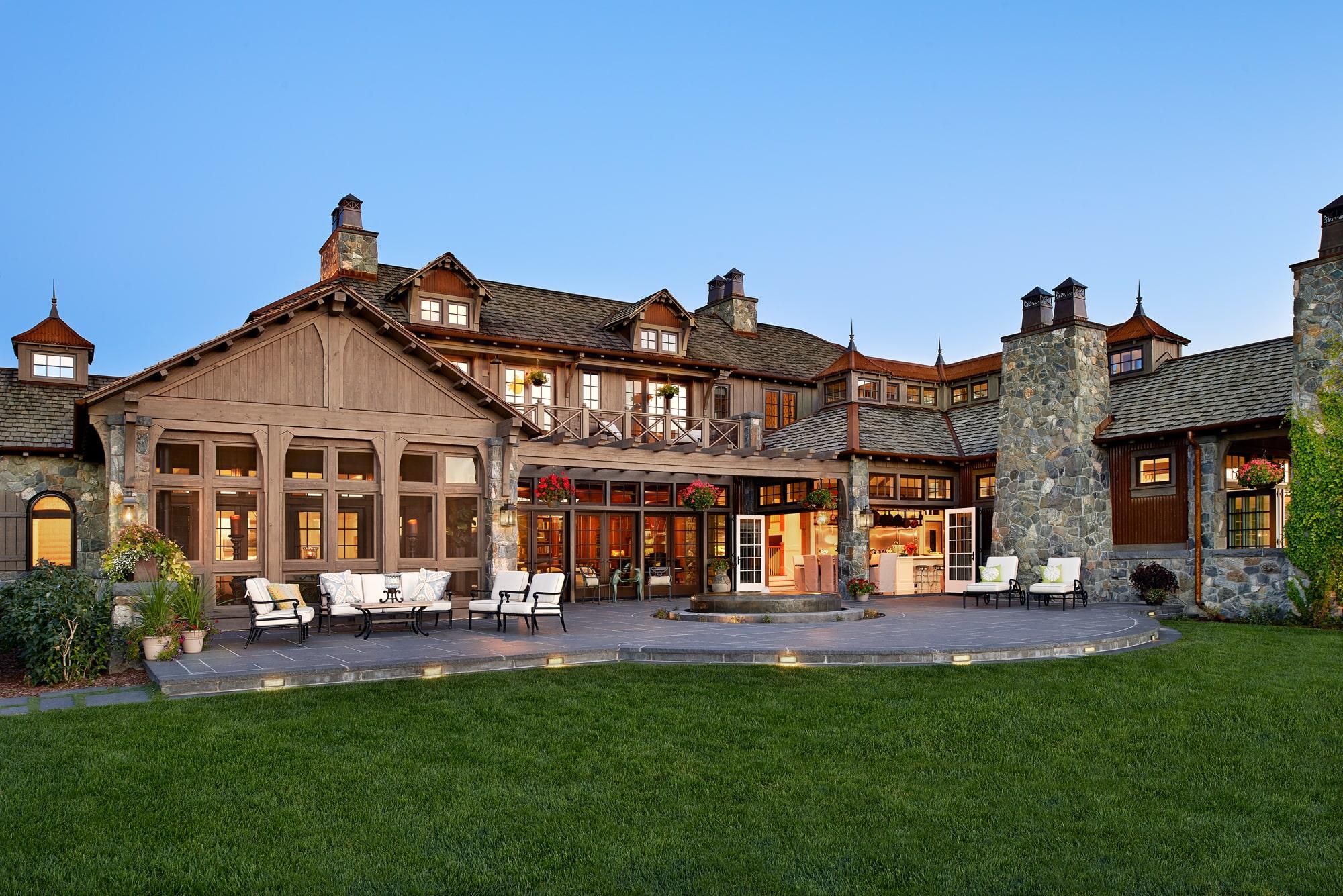 Large rustic-style estate with stone chimneys, expansive patio seating, and manicured lawn at twilight.