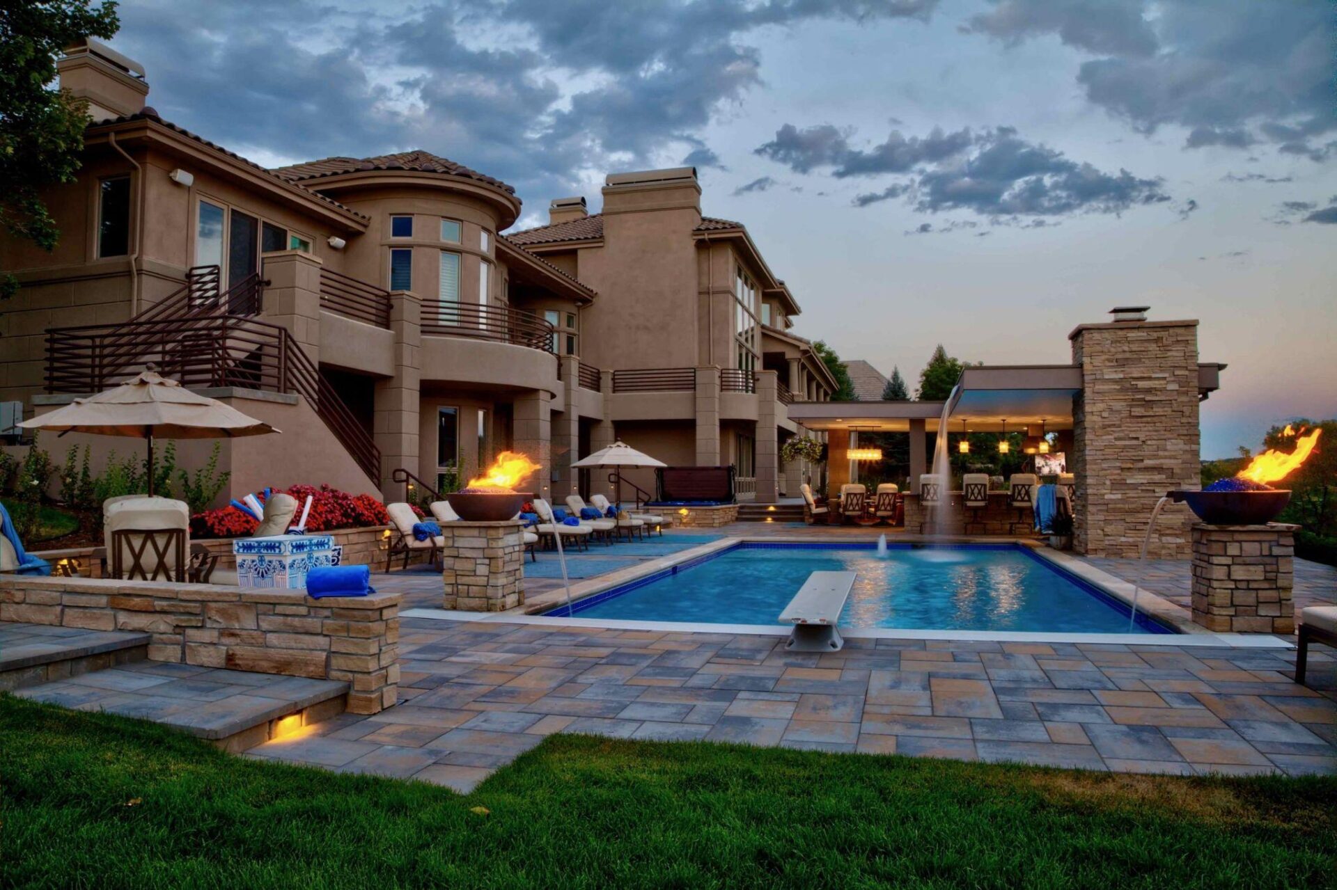 Luxury backyard featuring a rectangular swimming pool, stone fire bowls, lounge chairs with umbrellas, and a multi-level stucco home at dusk.