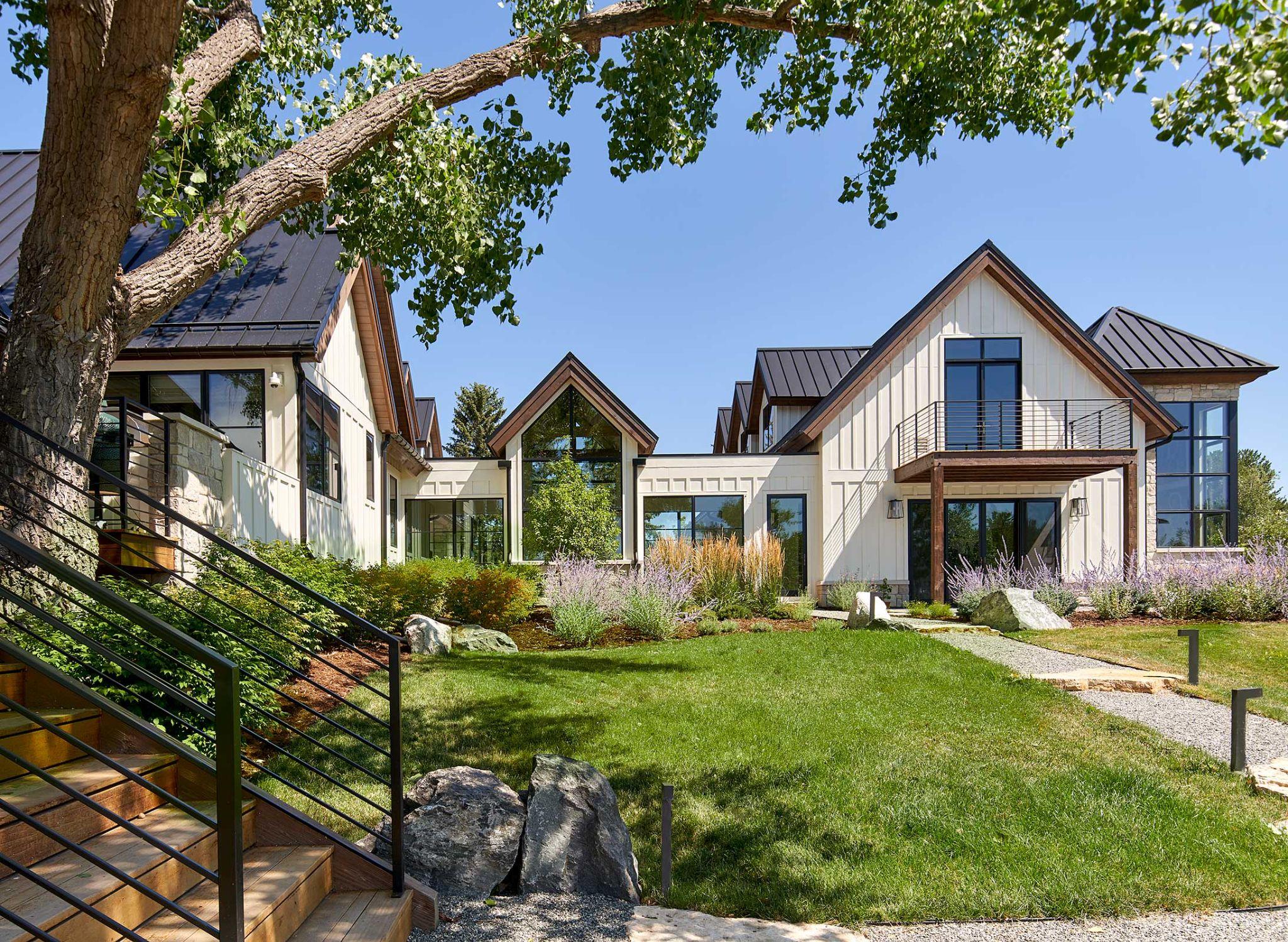 Modern farmhouse exterior with white siding, black metal roofs, landscaped gardens, and a central green lawn.