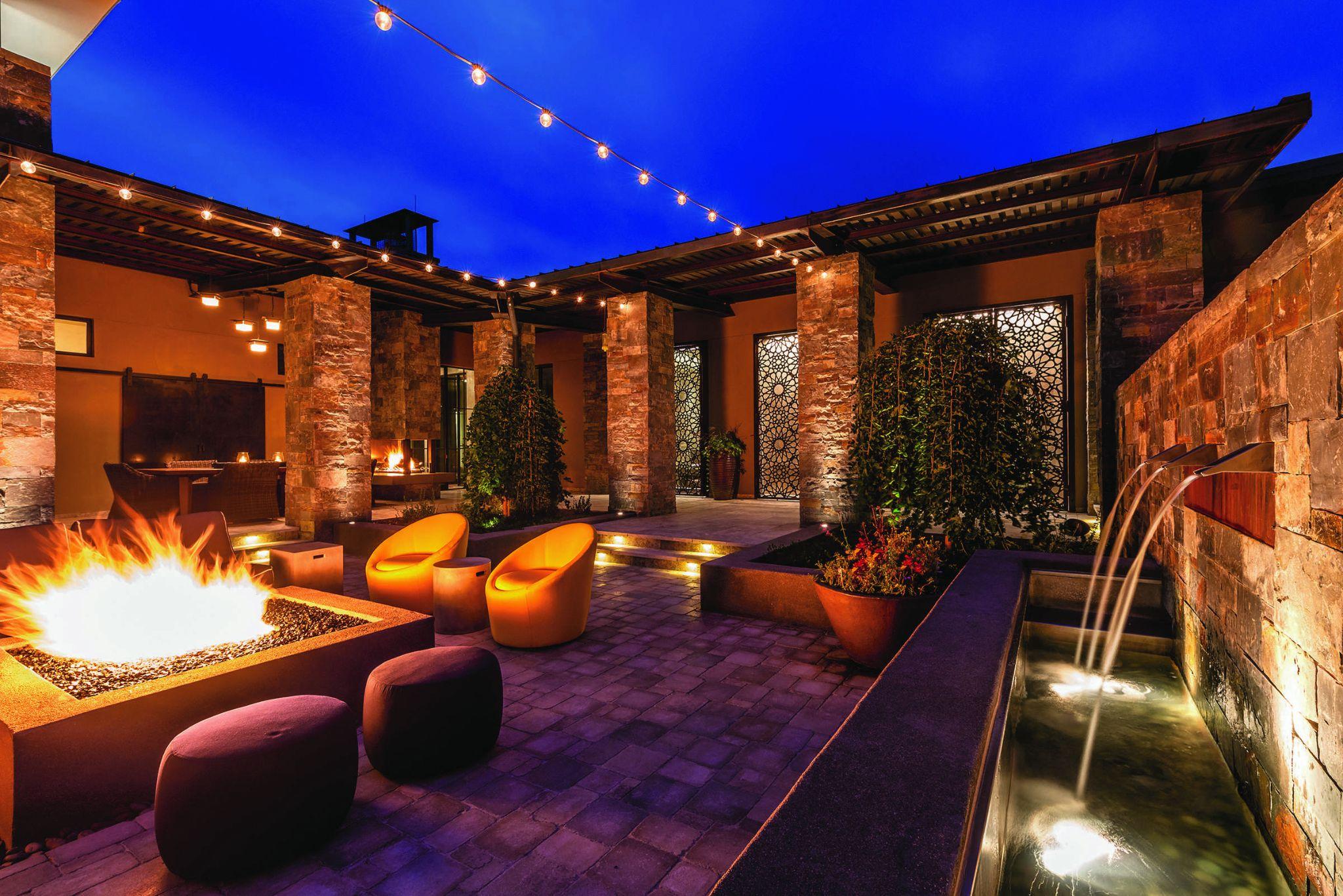 Modern outdoor patio at night with a large fire pit, string lights overhead, stone columns, water feature, and contemporary seating.