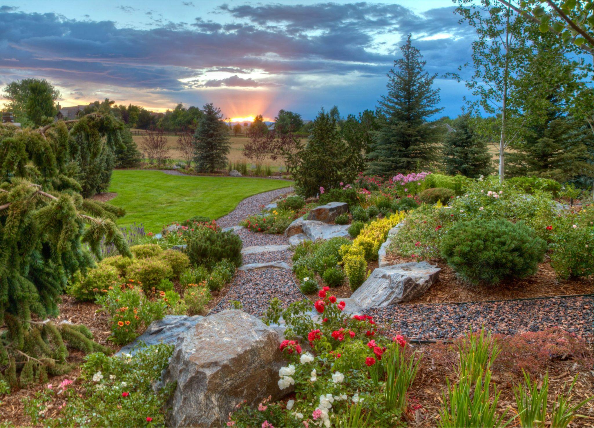 Naturalistic garden with winding stone paths, boulders, flowering plants, and evergreen trees under a dramatic sunset sky.