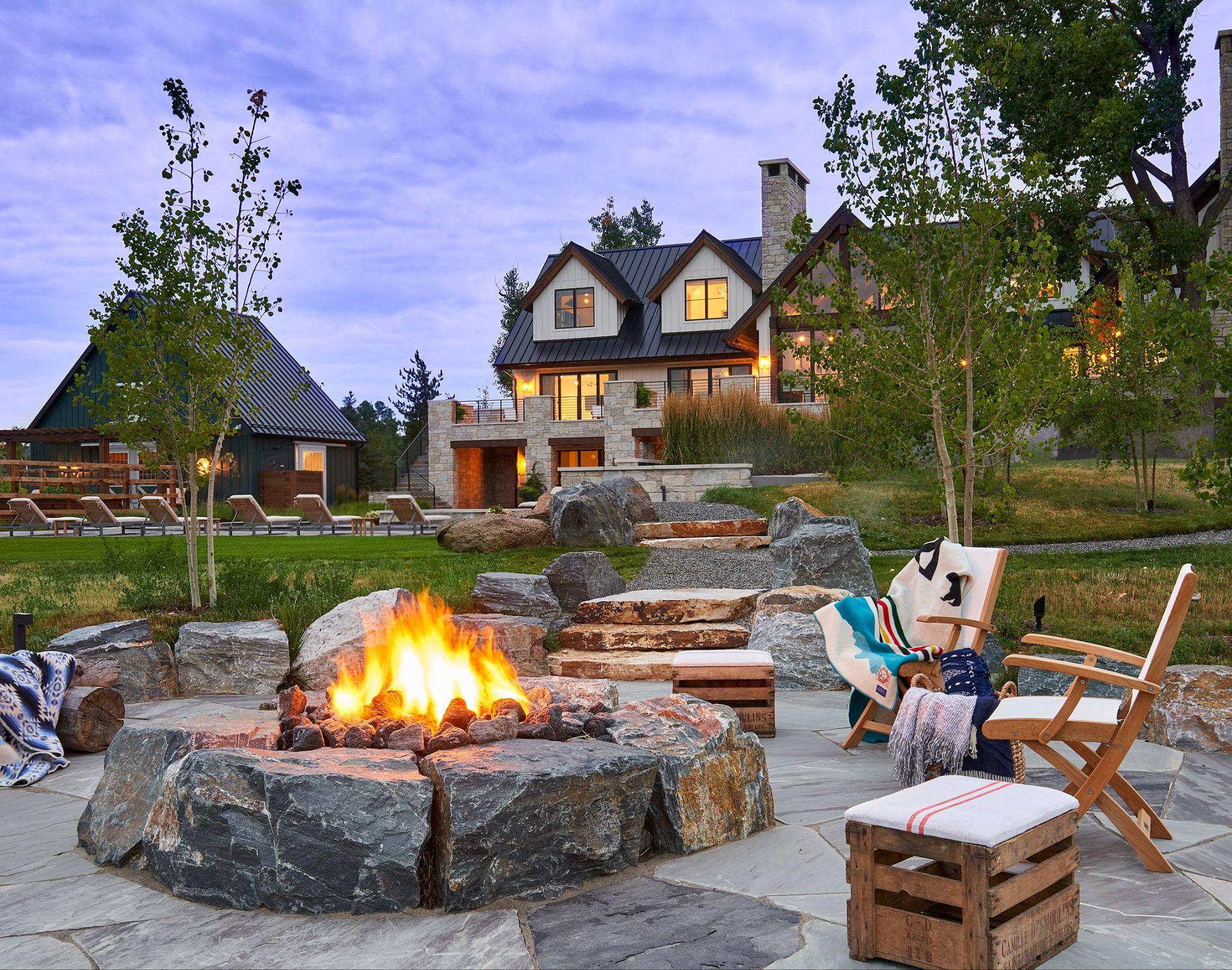 Outdoor stone fire pit surrounded by wooden chairs and blankets, set in a landscaped yard with a rustic modern home in the background.