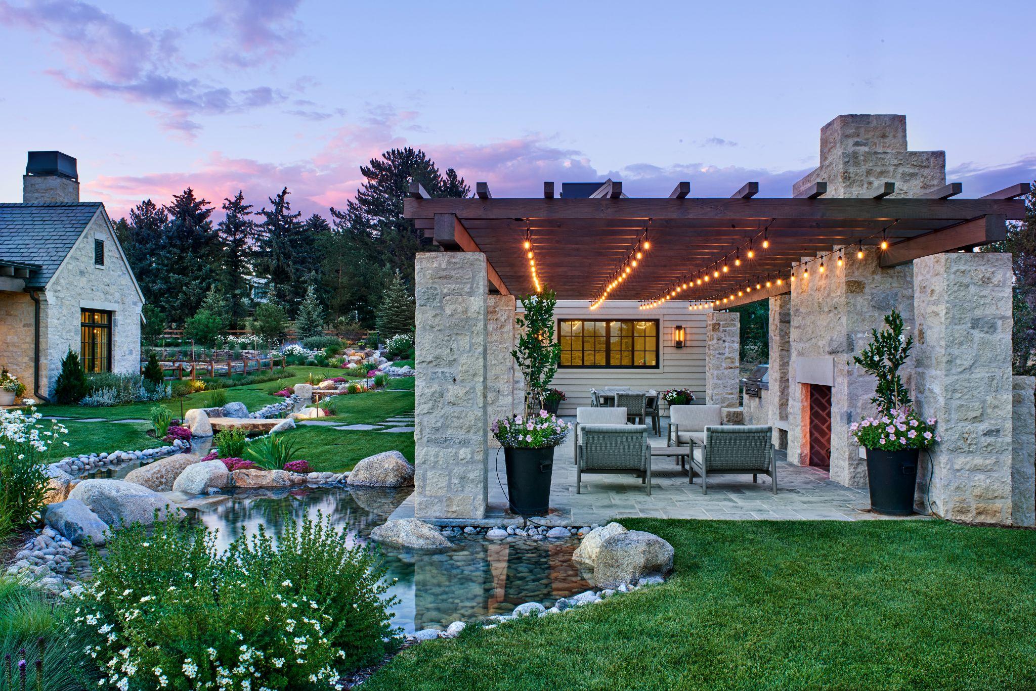Pergola-covered outdoor seating area with string lights, stone fireplace, and reflective pond surrounded by lush landscaping.