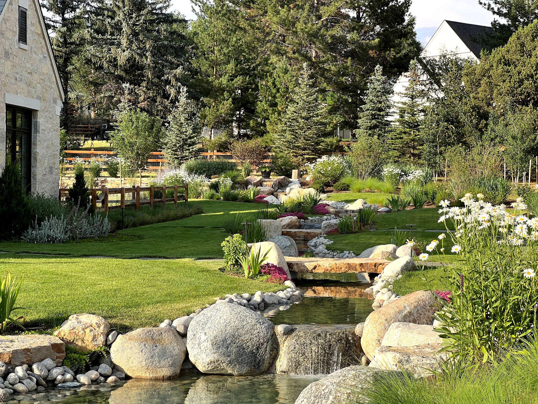 Tranquil backyard stream with small waterfalls flowing through stone-lined channels and manicured lawn plantings.